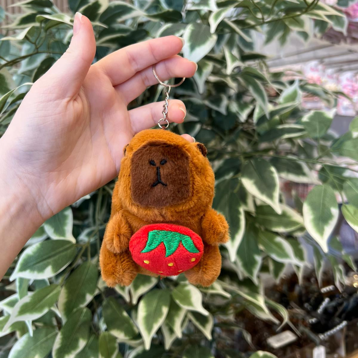 A hand holds a small, brown, stuffed animal bear keychain that has a strawberry on its belly. The background is leafy and blurred.