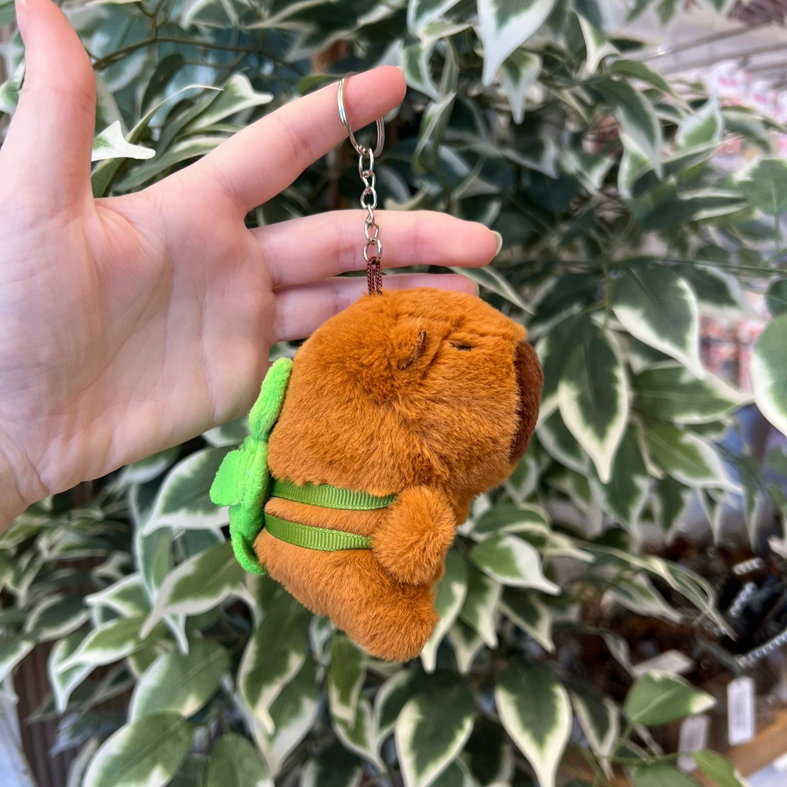 A hand holds a brown plush capybara keychain with a green leaf detail against a background of green and white leaves.