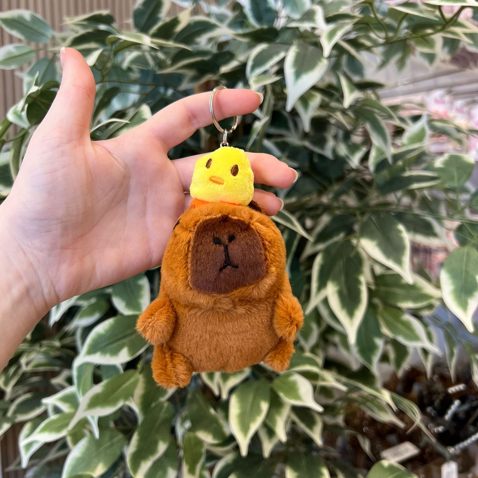 A hand holds a brown plush bear keychain with a yellow chick on its head, against a leafy green background.