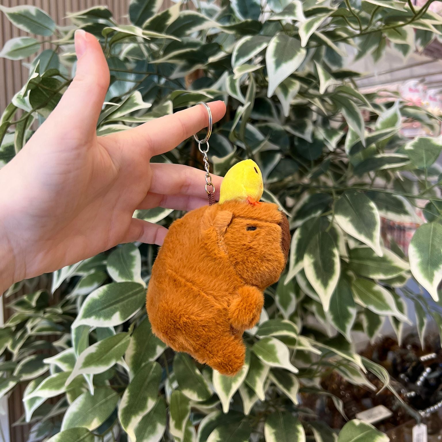 A hand holds a keychain featuring a brown, plush animal figure with a yellow bird perched on its back, set against a backdrop of variegated green and white leaves.