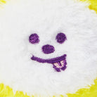 Close-up of a white, plush toy with a simple embroidered purple face. The toy has yellow accents around the edges. The face consists of two circular eyes, a nose, and a smiling mouth with a tongue sticking out.