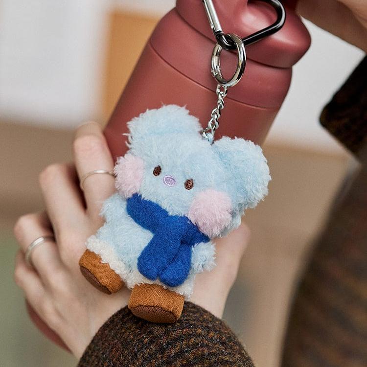 Close-up of a blue plush bear keychain with pink cheeks and a blue scarf, attached to a maroon water bottle with a metal carabiner. The keychain is held in a hand with two silver rings.