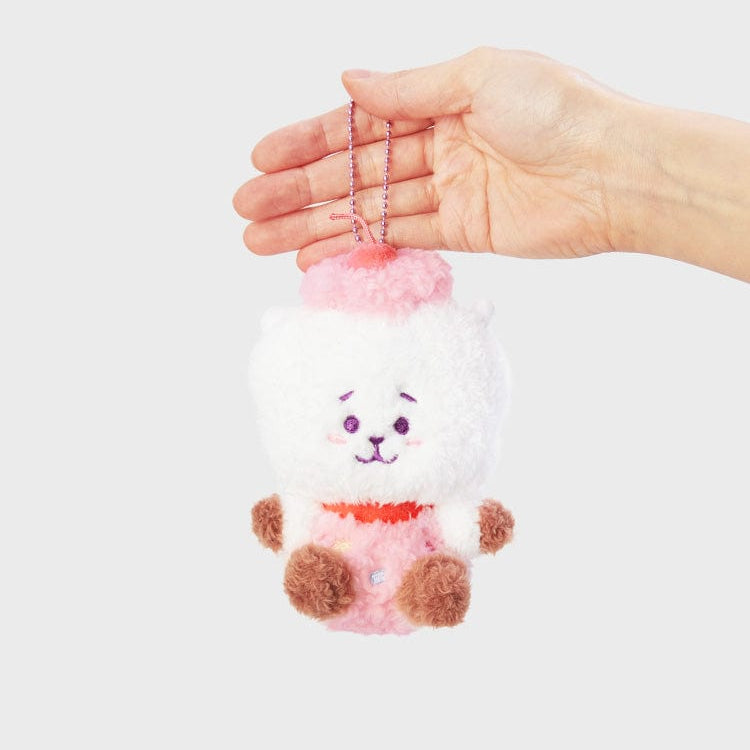 Close-up of a stuffed animal keychain featuring a white, fluffy bear with a pink cherry on its head, held up by a fair-skinned hand against a gray background.