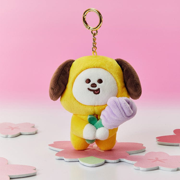 Yellow plush Chimmy keychain holding a purple flower, standing on a pink cherry blossom coaster, with pink background.