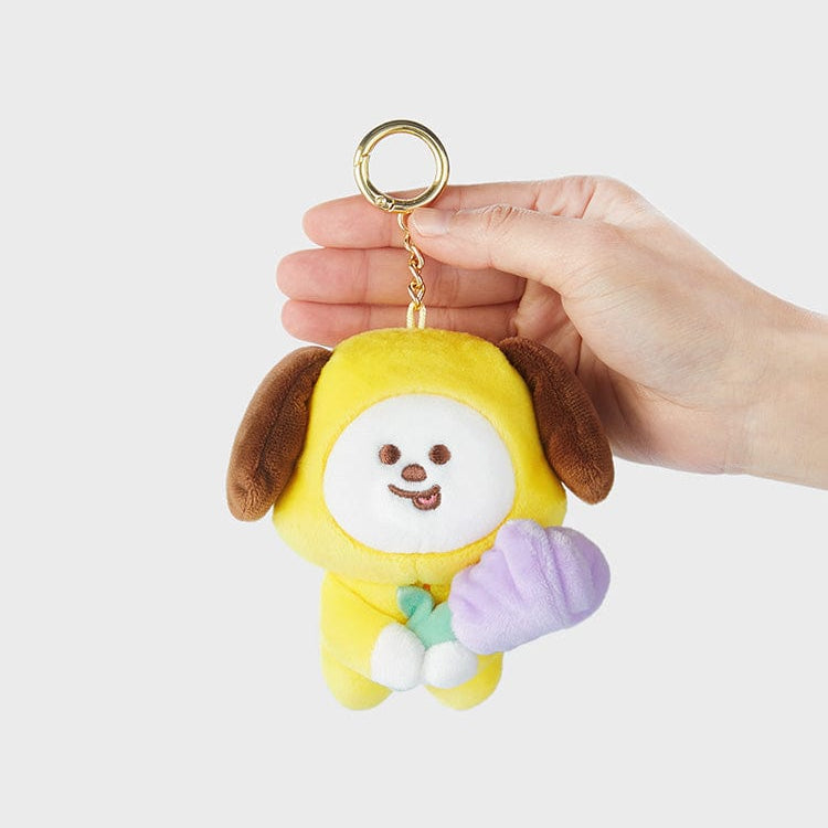 A yellow, plush Chimmy keychain with brown ears is held by a hand. Chimmy is holding a purple flower.
