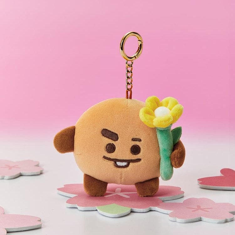 Brown plush toy keychain of a cartoon character holding a yellow flower, standing on a flower-shaped coaster against a pink background.