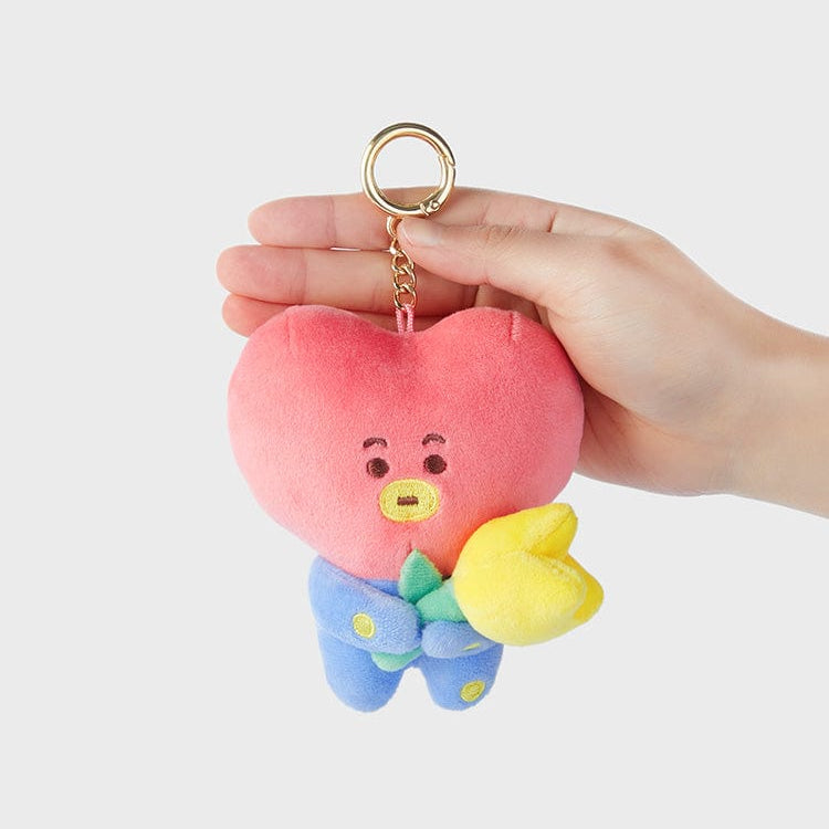 Hand holding plush pink heart-shaped character keychain with blue outfit and yellow flower.
