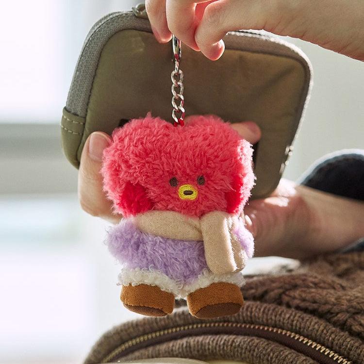 Close-up of a TATA plush keychain hanging from a khaki pouch. The plush is pink, purple, and brown with a beige scarf. The keychain has a silver chain. The background is blurred.