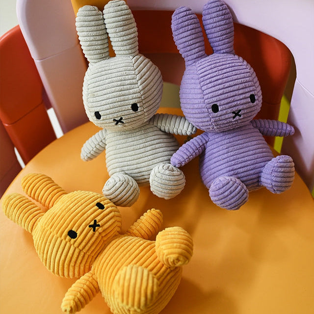 Three plush bunny toys in yellow, gray, and purple, arranged on an orange chair. Each toy has a corduroy texture and simple stitched facial features.