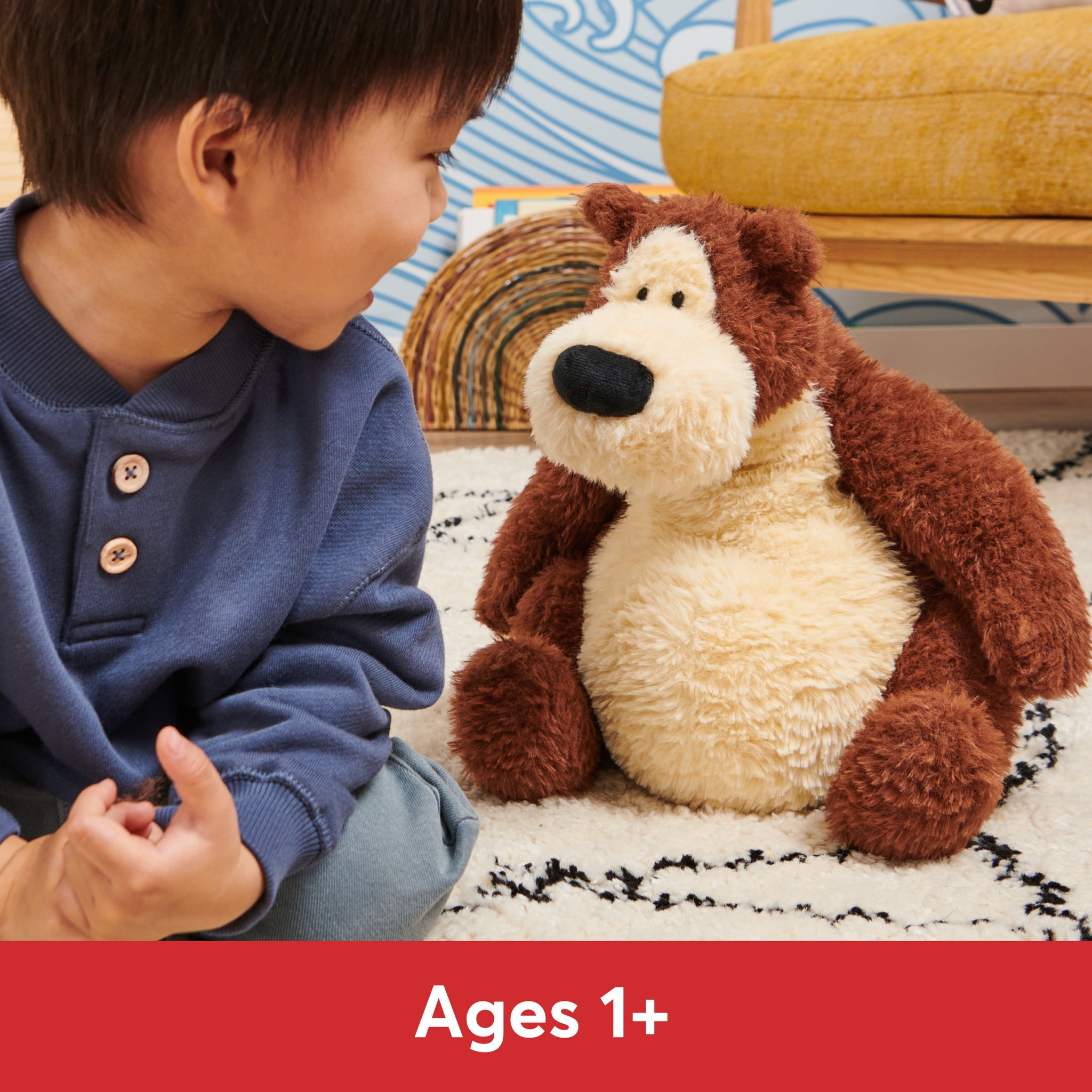 A young boy sits on a cream rug with black accents, gazing at a brown and tan stuffed bear. The boy wears a dark blue button-up shirt and light blue pants.