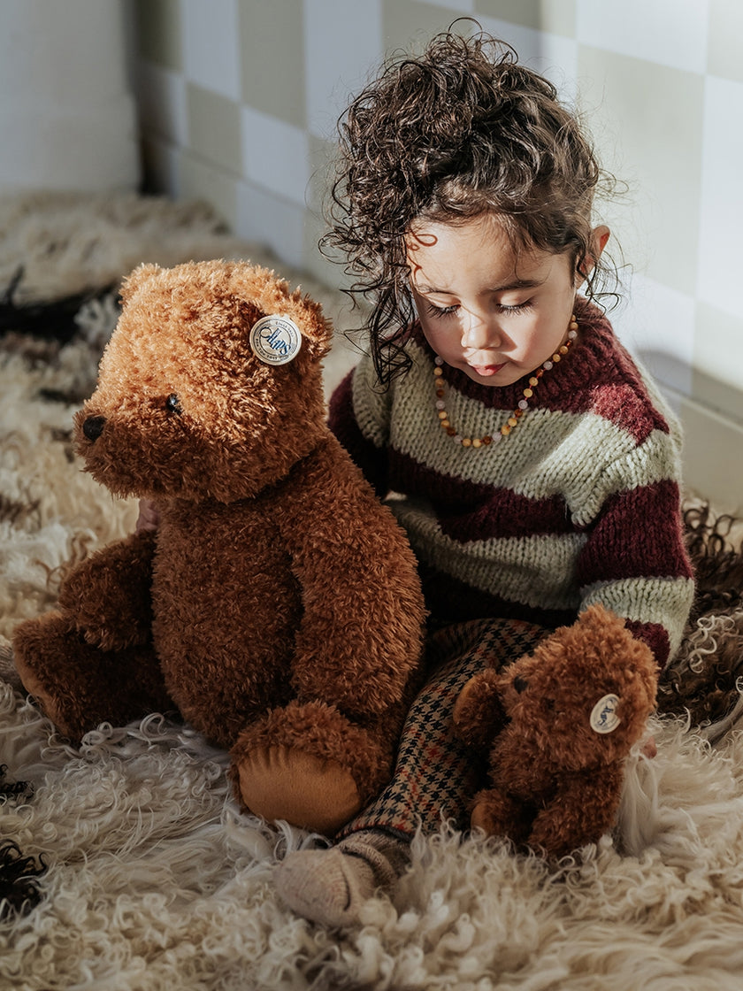 Child with curly hair wearing a striped sweater and amber necklace, seated with two teddy bears on a shaggy rug.