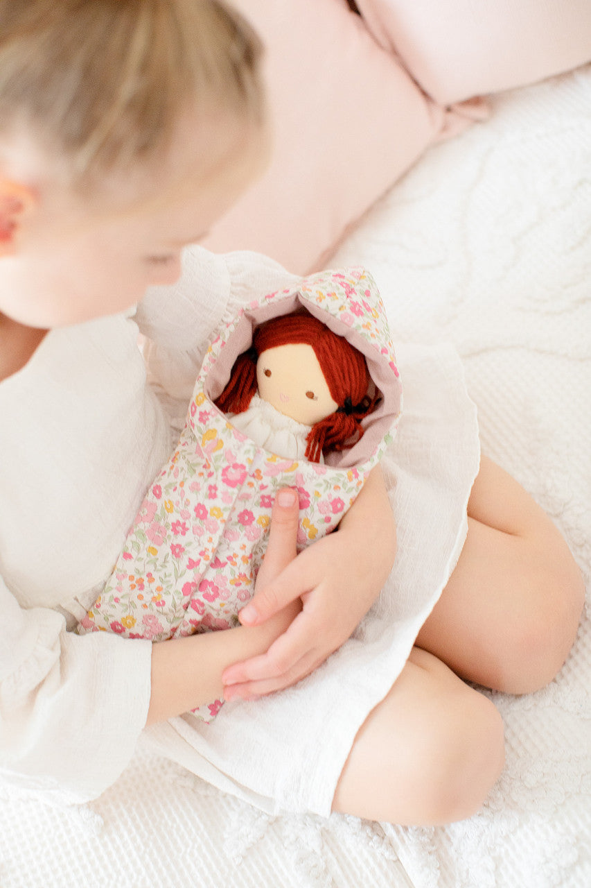A young child in a white dress sits and holds a doll wrapped in floral fabric.