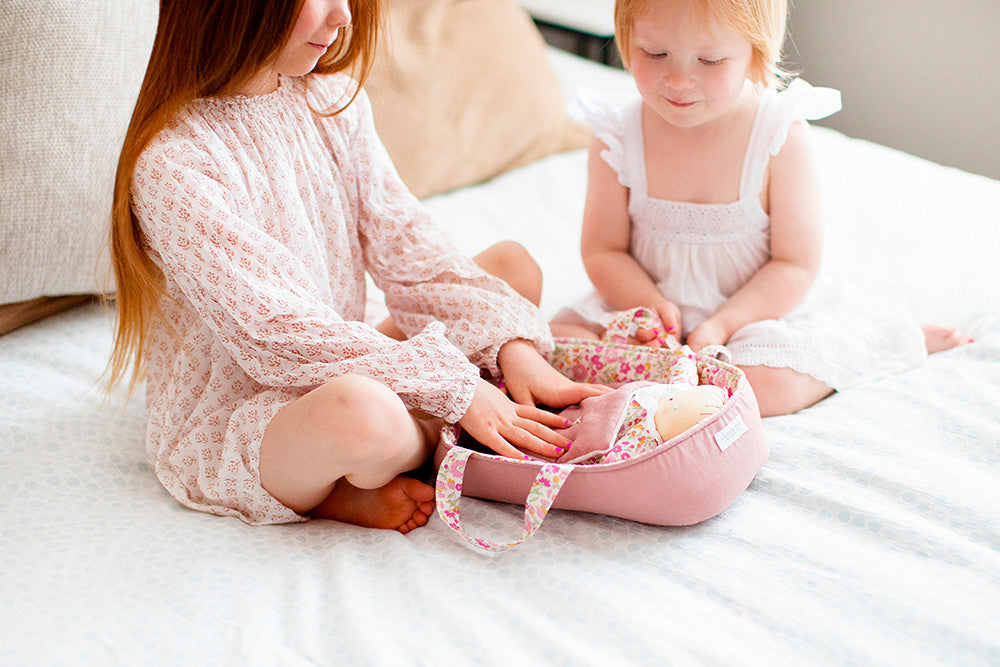 Two red-haired girls sitting on a bed with a pink doll carrier and doll.