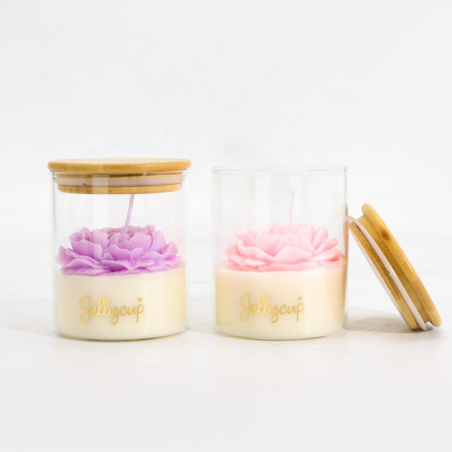 Two clear glass jar candles with bamboo lids on a white background. One candle has a purple flower-shaped wax top, and the other has a pink flower-shaped wax top.