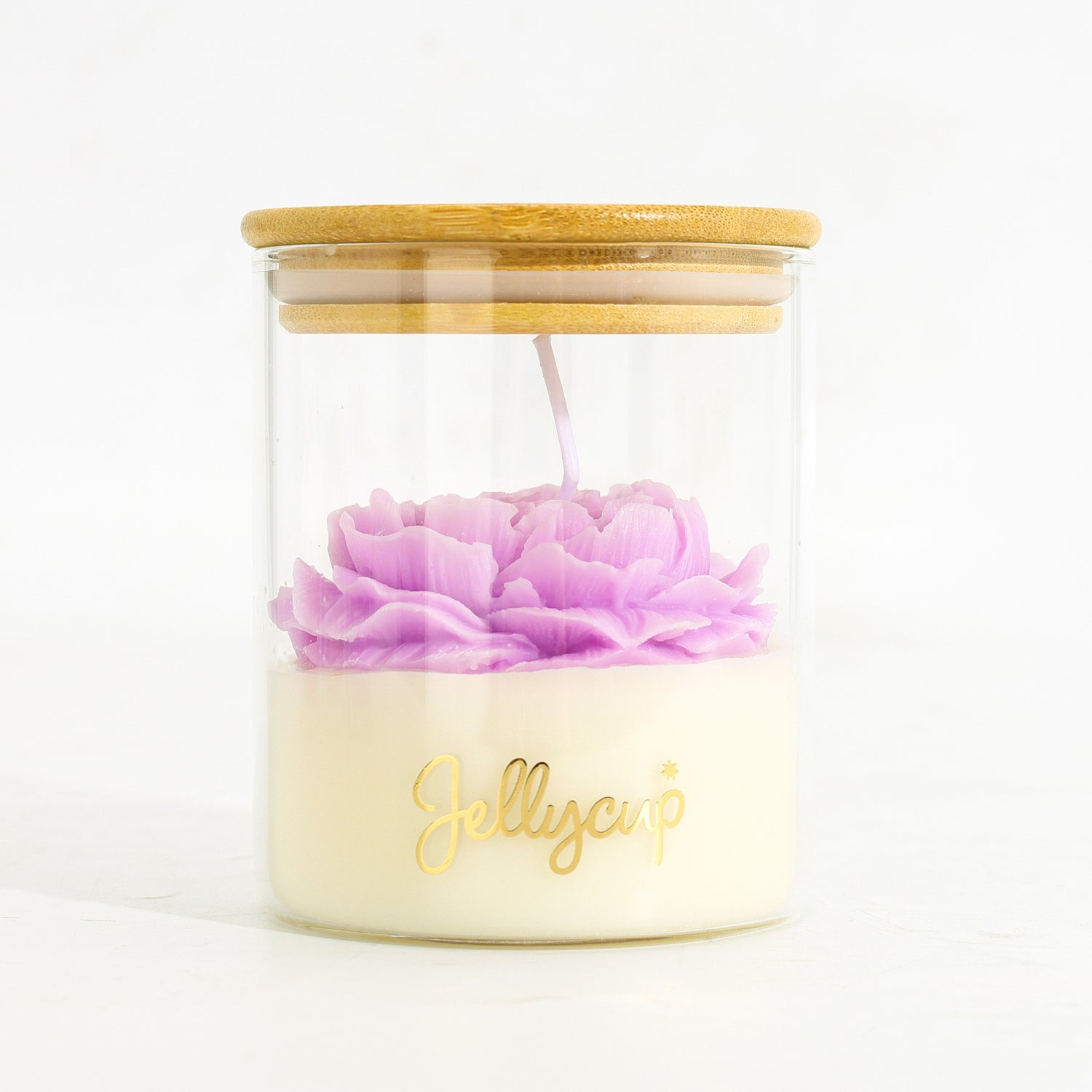 A clear glass jar candle with a light wood lid, filled with creamy off-white wax and topped with a sculpted light purple flower. The word 'Jellycup' is printed in gold on the front of the jar.