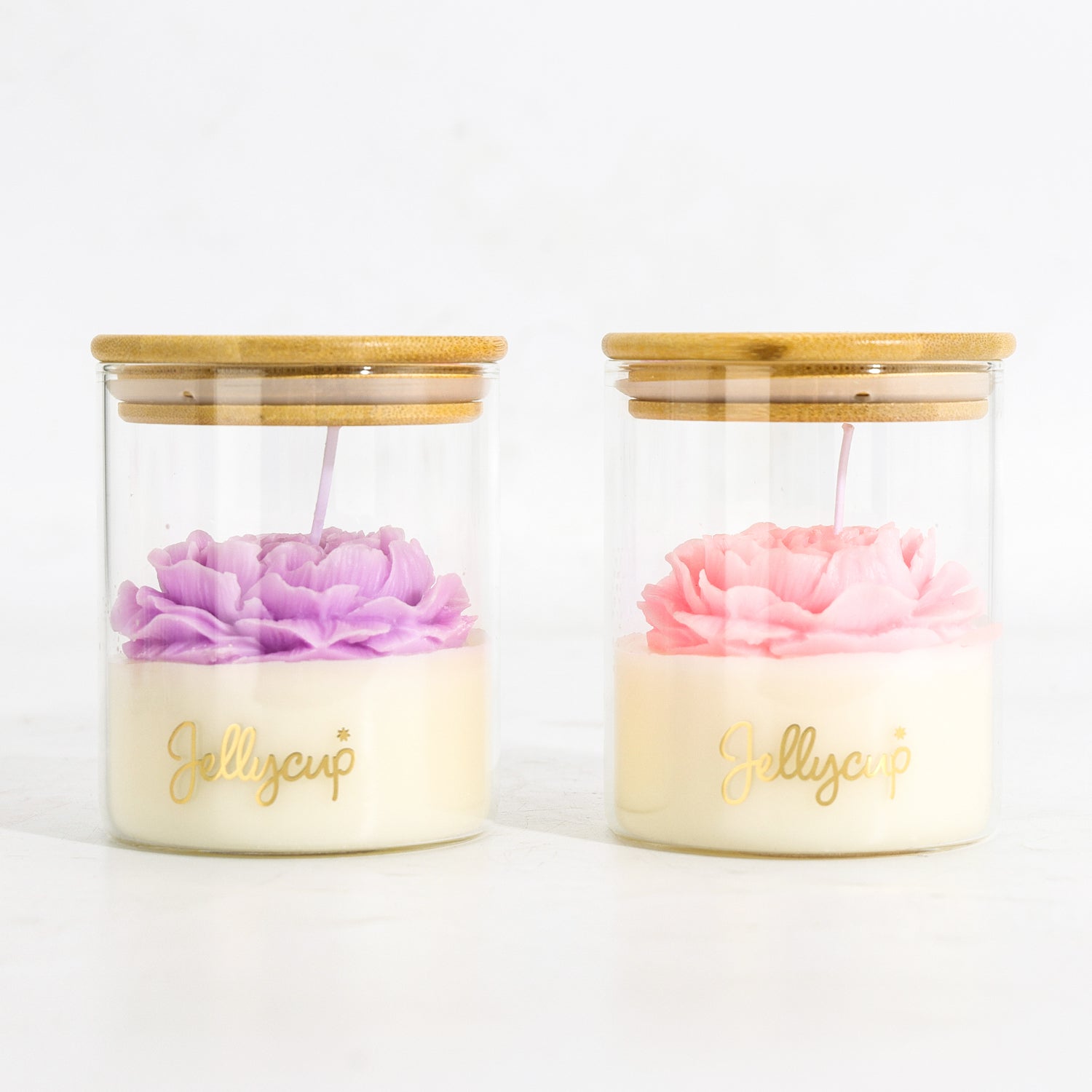 Two glass jar candles with bamboo lids on a white background. The left candle has a purple flower and the right candle has a pink flower, both resting on a cream-colored wax base with gold 'Jellycup' branding.