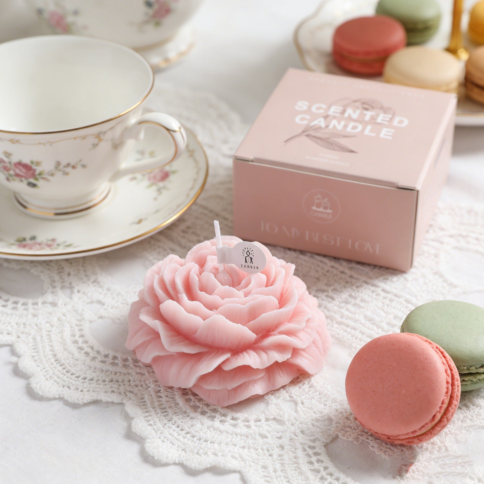 Pink peony candle by Jellycup with a white label on a lace doily, surrounded by macarons and tea cup.