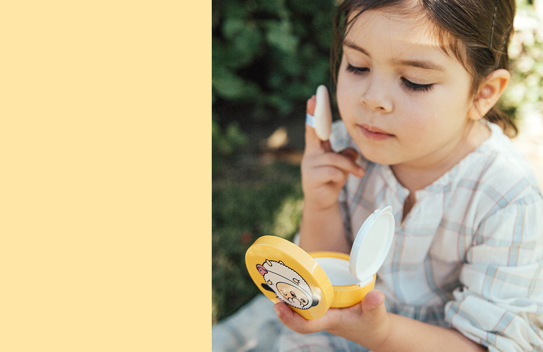 Young child applying Puttisu Sun Cushion outdoors using the included puff applicator. The bright yellow compact features a cute character design and is held open in her hand.