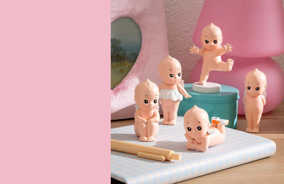 Collection of Smoko Kewpie blind box figurines displayed on a tabletop, featuring small baby-like characters in playful poses. The pastel background and props highlight the cute and collectible design of the figures.
