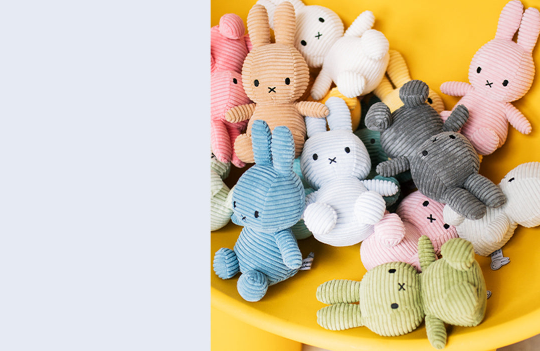 Colorful collection of Bon Ton Toy Miffy plush bunnies made from soft corduroy fabric in pastel and neutral shades, displayed on a yellow background.