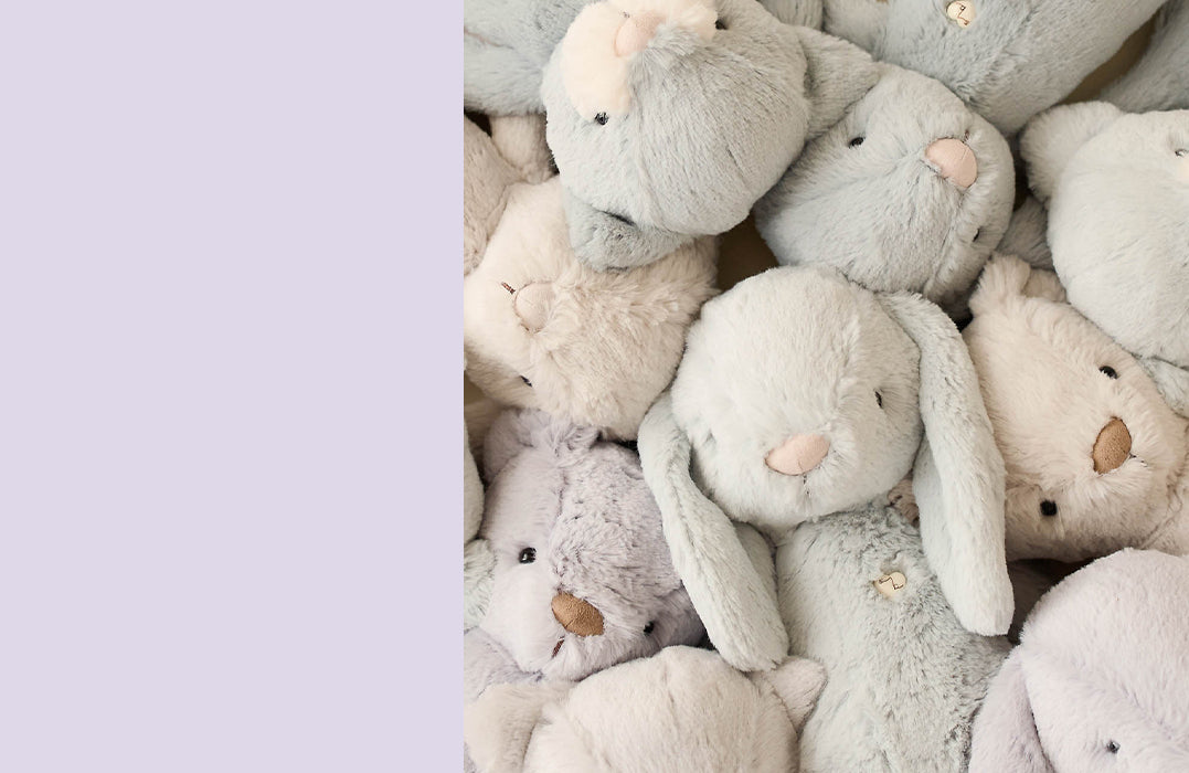 A pile of Snuggle Bunnies plush toys in soft pastel shades of grey, cream, and lavender, featuring floppy ears and embroidered noses. These cuddly stuffed animals are designed for comfort, making them perfect snuggle companions and thoughtful gifts.