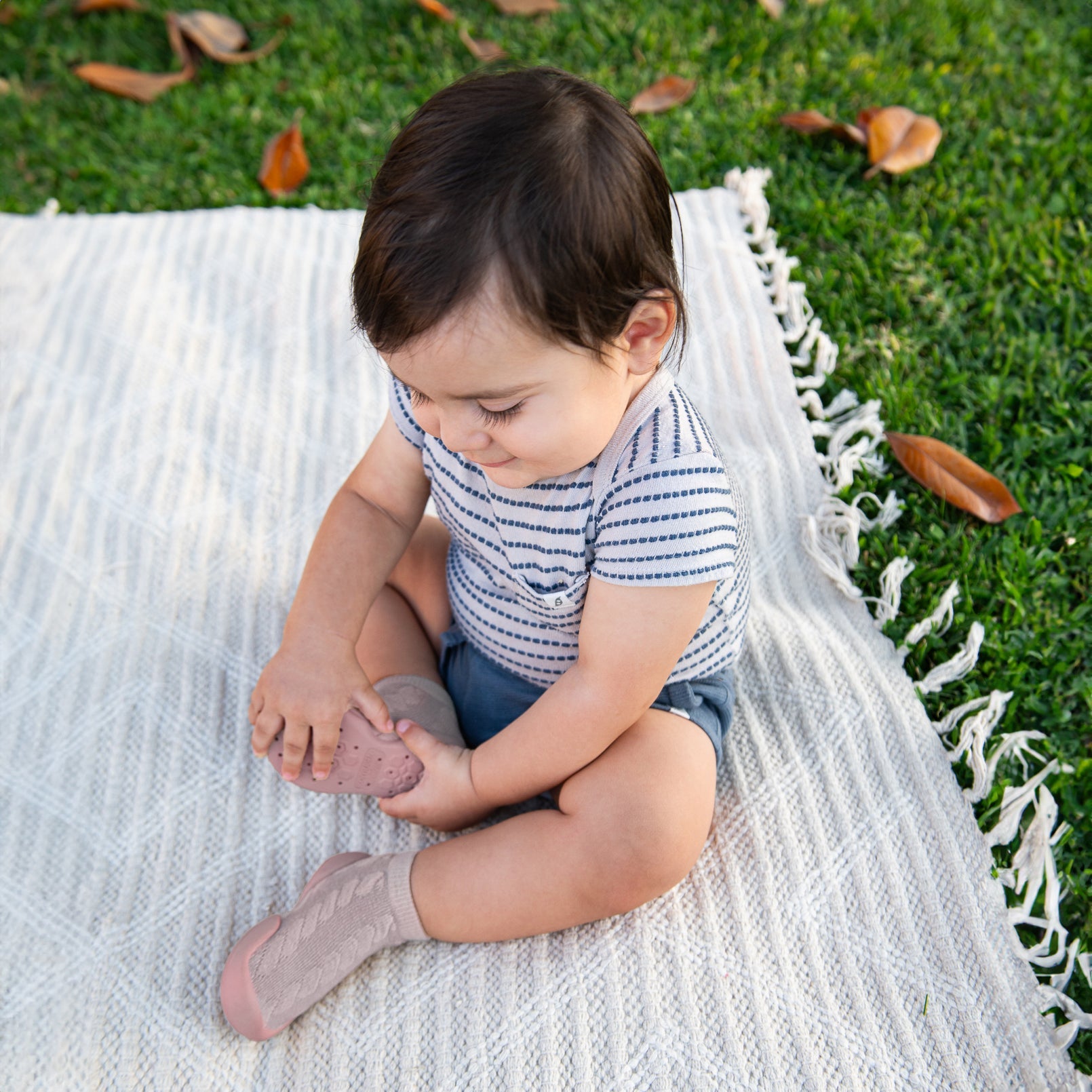 A baby sitting on a white blanket wearing a striped shirt, blue shorts, and pink socks.