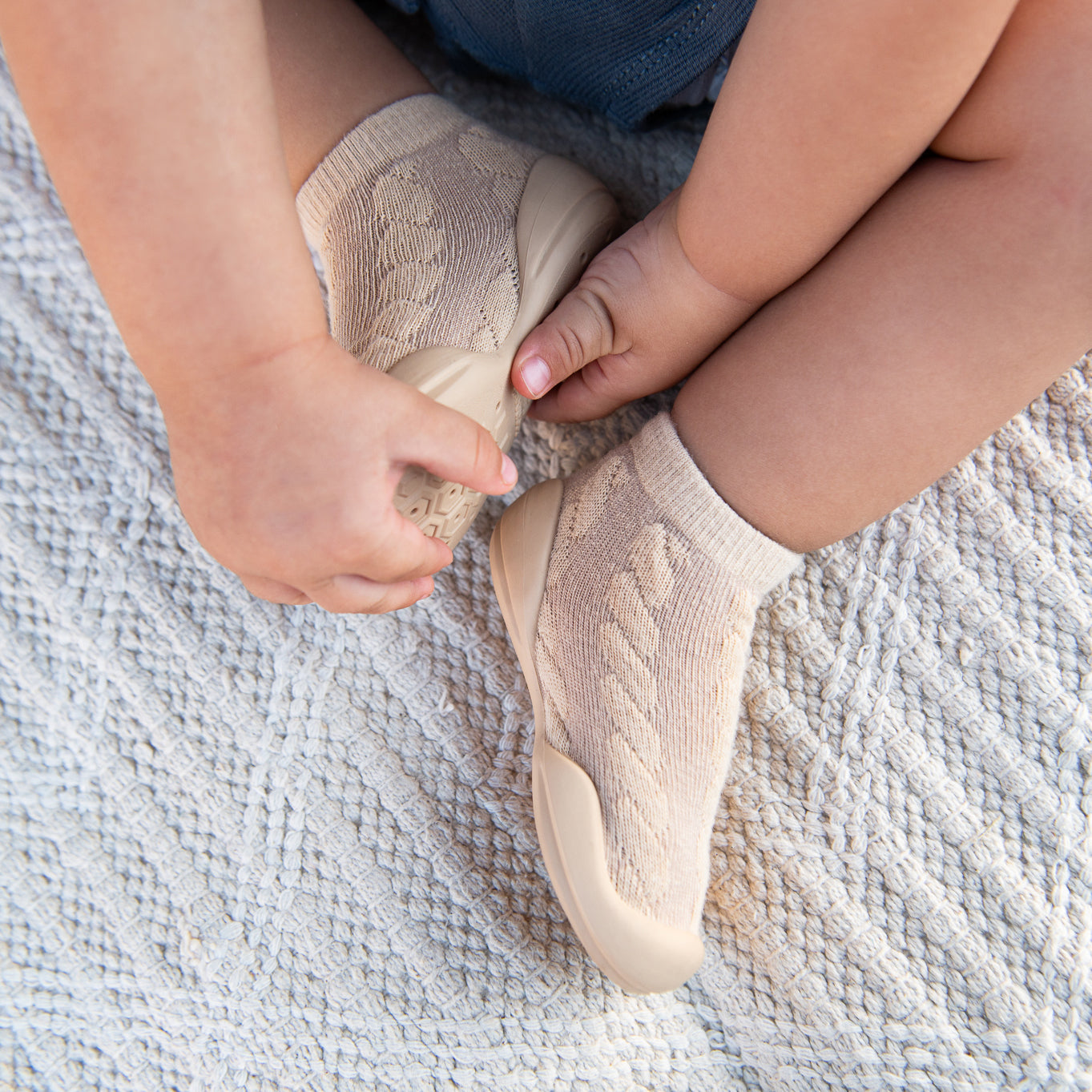 Close-up of a baby wearing sock shoes, sitting on a textured cream blanket.