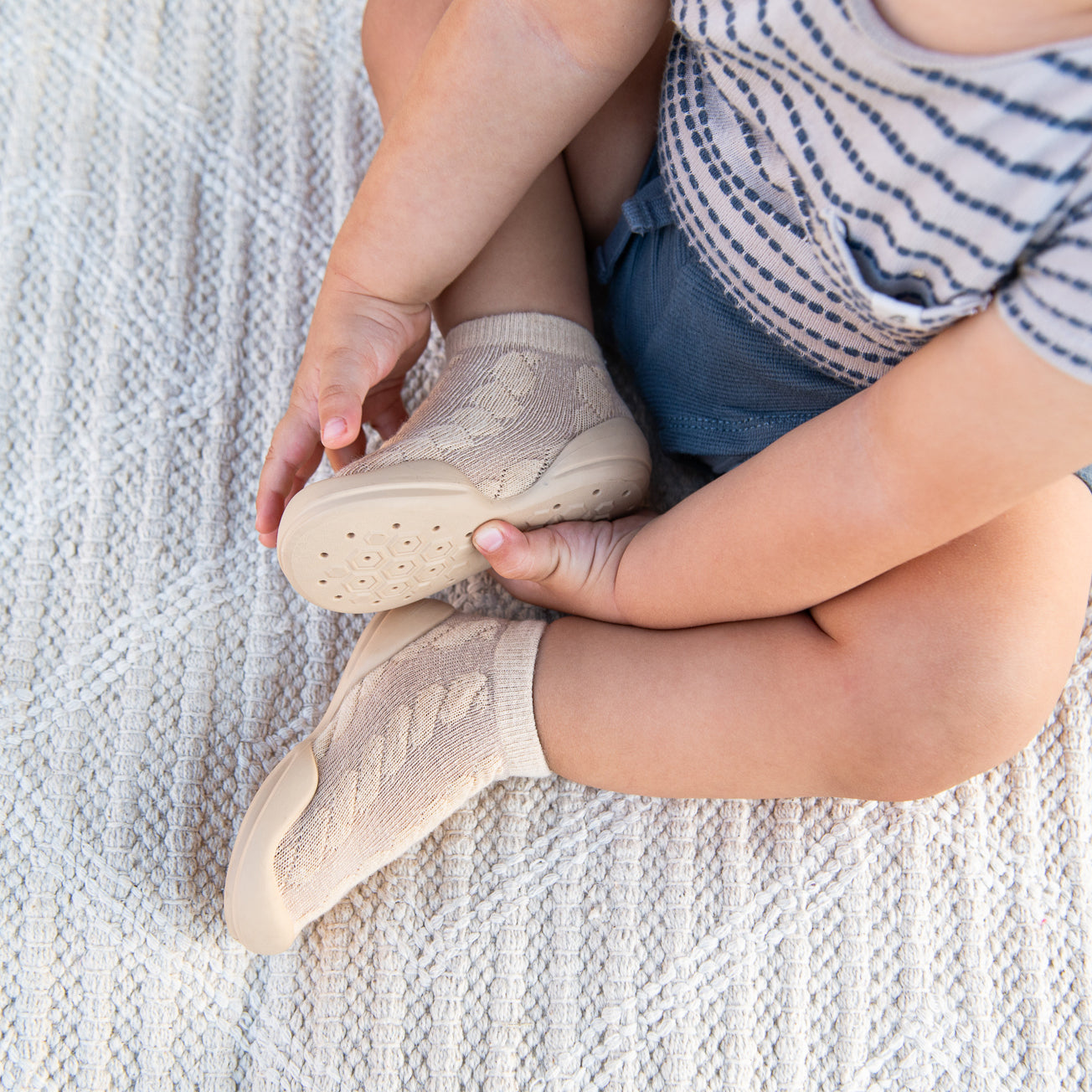 A baby wearing tan sock-shoes sitting on a patterned blanket.