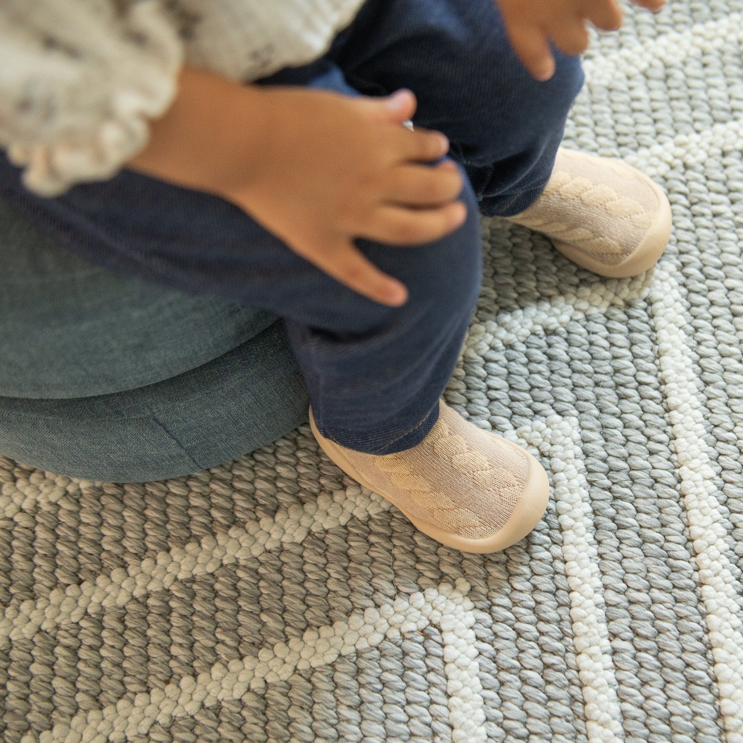 Child wearing blue pants and light brown shoes sitting on a blue cushion on a gray and white rug.