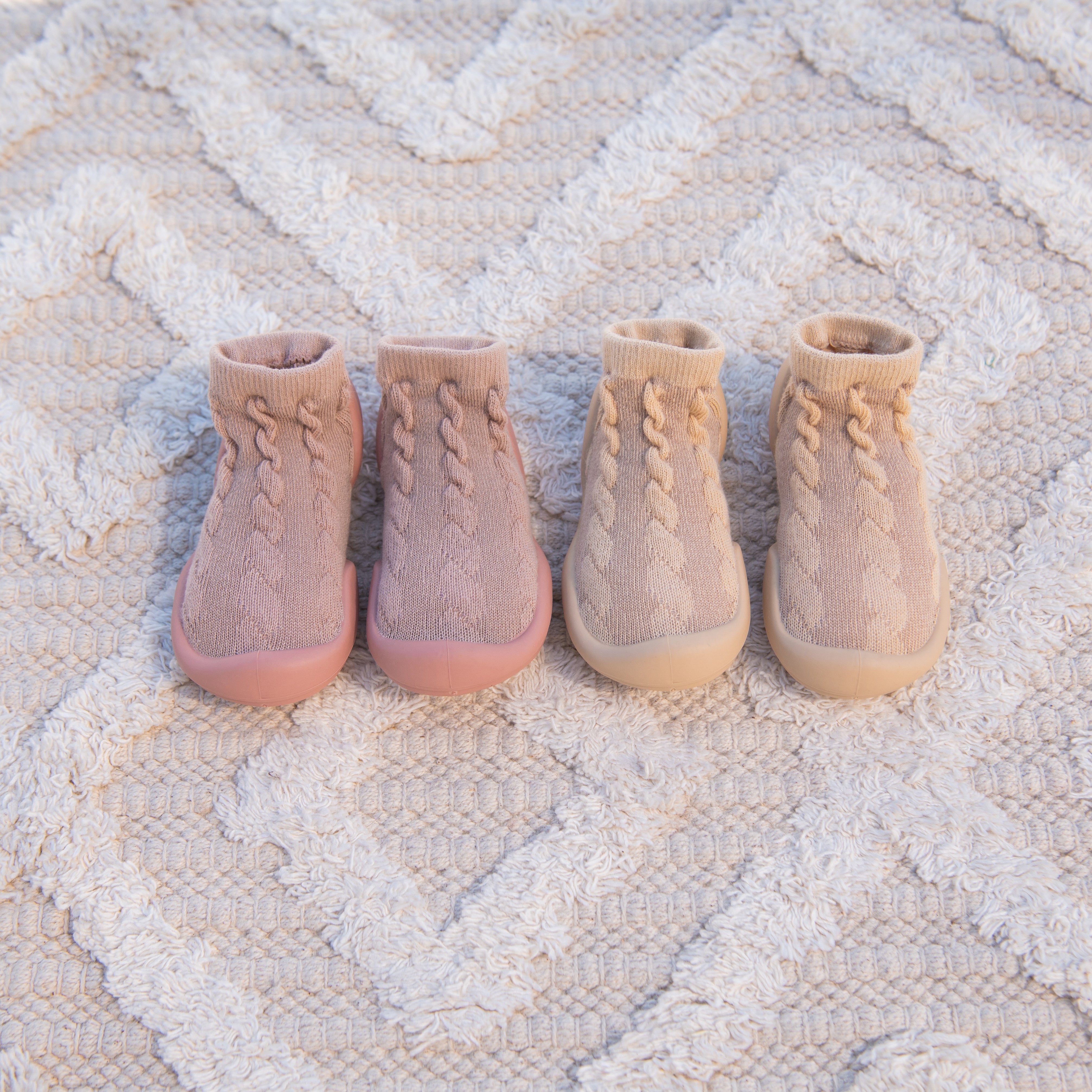 Four baby socks with rubber soles in varying shades of beige and pink on a white textured rug.