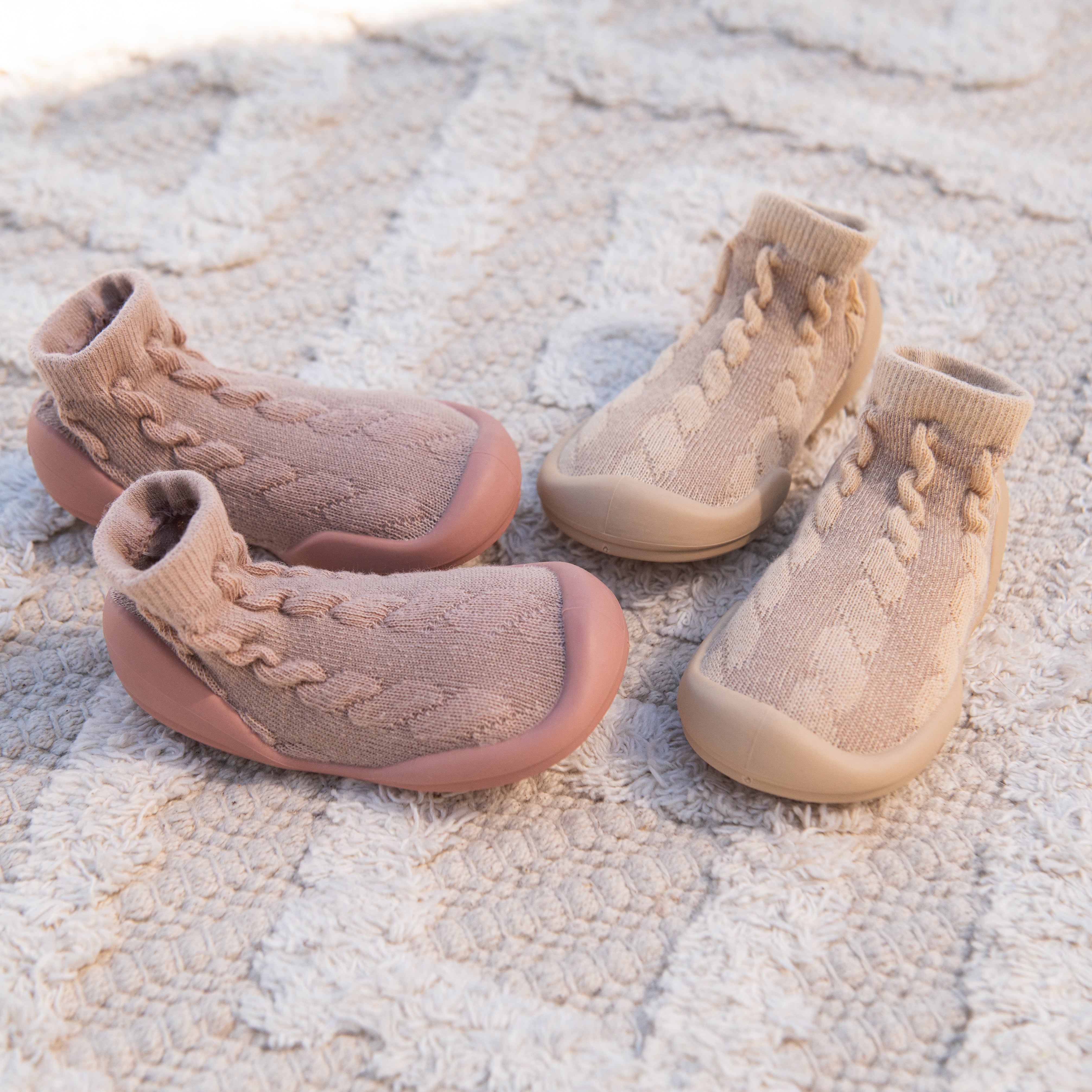 Two pairs of baby sock-shoes on textured white fabric. One pair is pink, the other is beige.