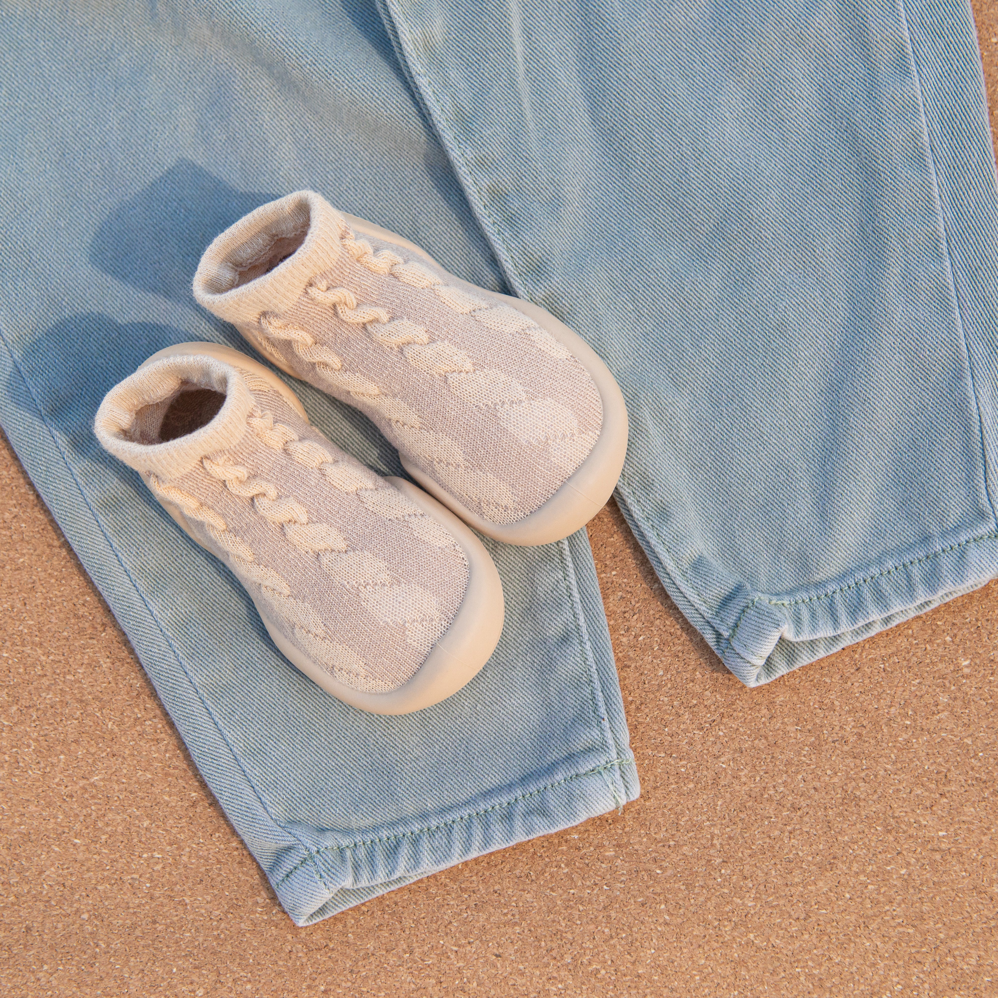 Two neutral baby sock shoes on light blue denim on a cork surface.