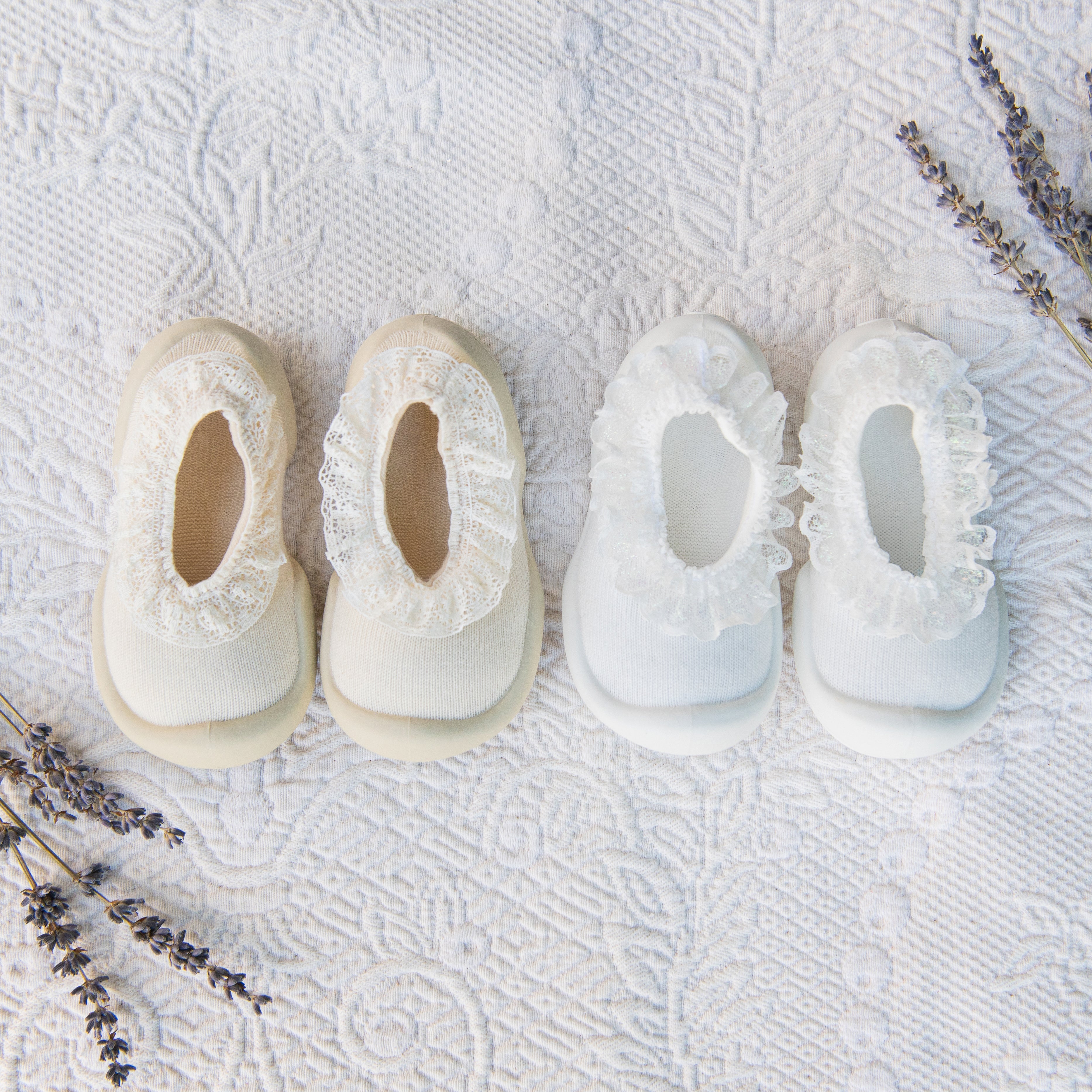 Two pairs of baby shoes, one cream and one white, with a ruffled trim, sit on a textured white fabric with sprigs of lavender.