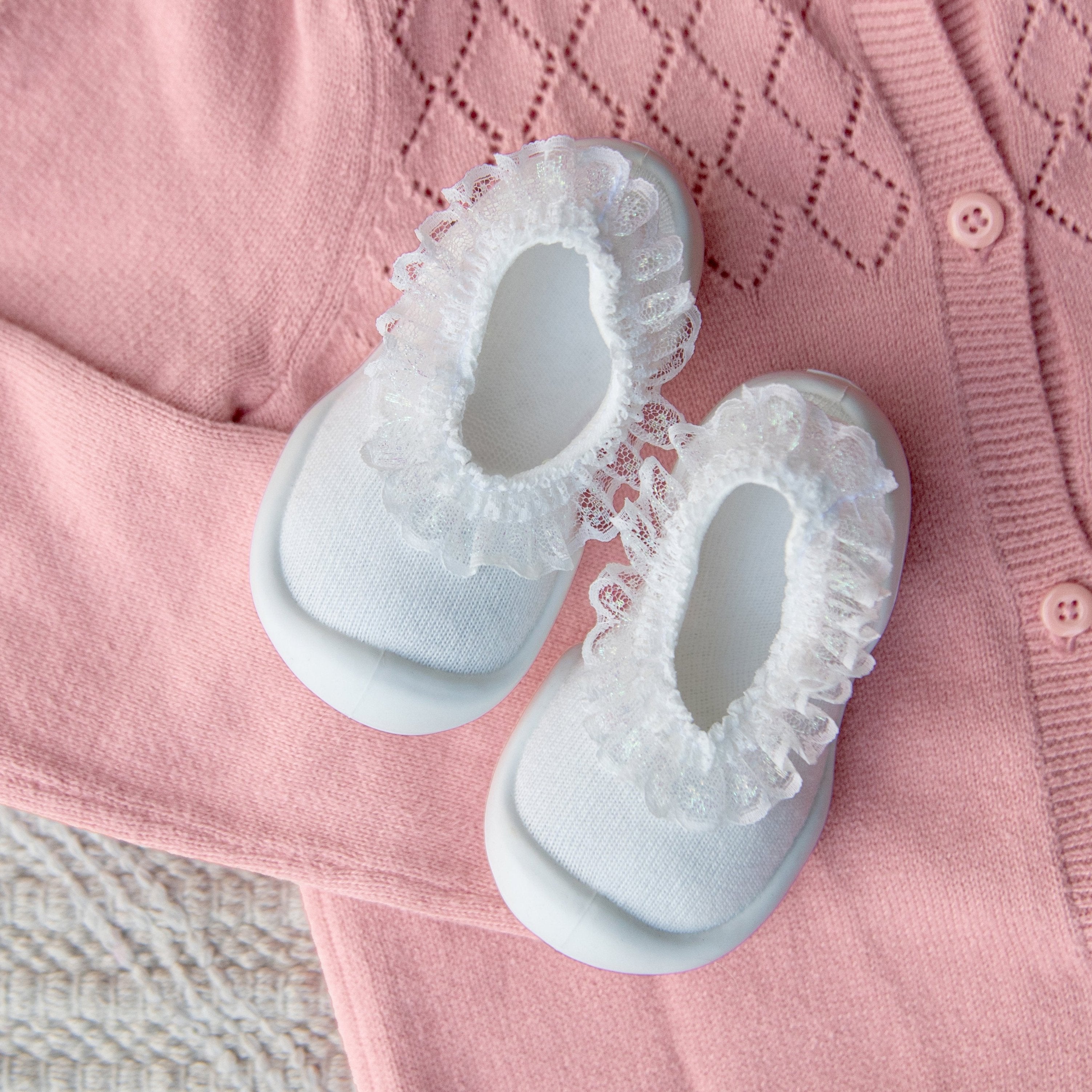 Two white baby shoes with lace trim on a pink knit cardigan.