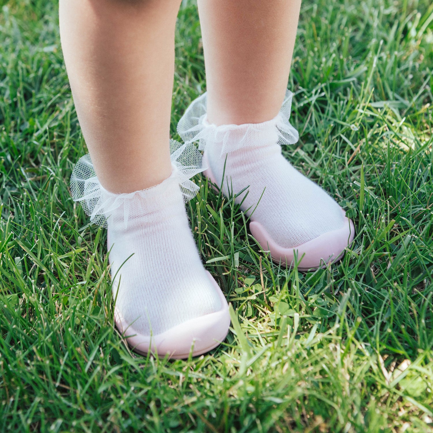 Baby legs with white frilly socks and pink-soled shoes on green grass.