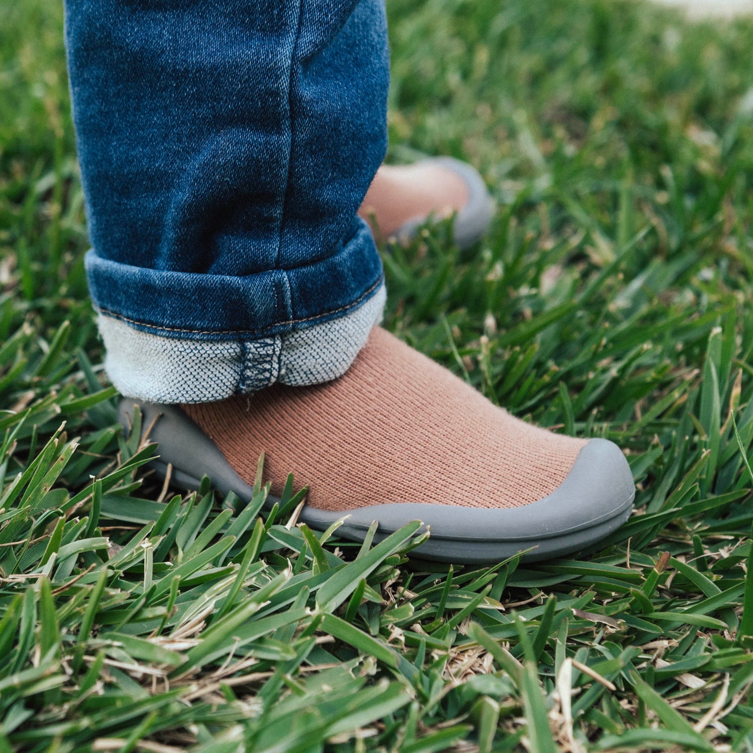 Person standing in grass, wearing blue jeans with cuffs and brown shoes with gray soles.