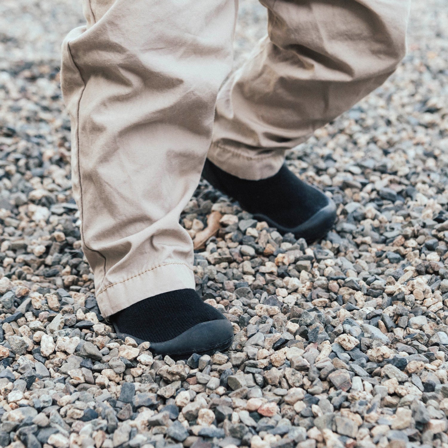 Person wearing black sock shoes and beige pants standing on gravel.