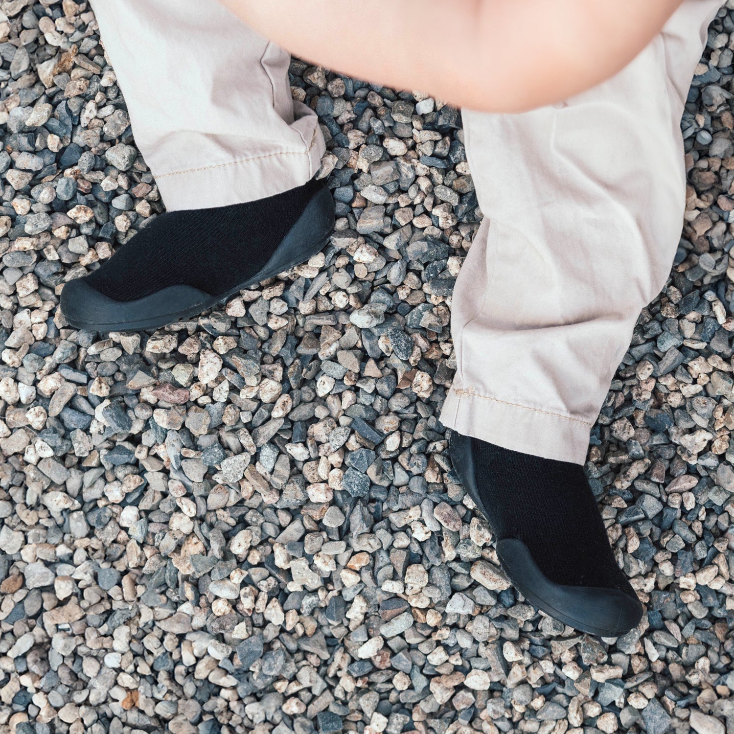 Person wearing tan pants and black sock shoes on a gravel ground.