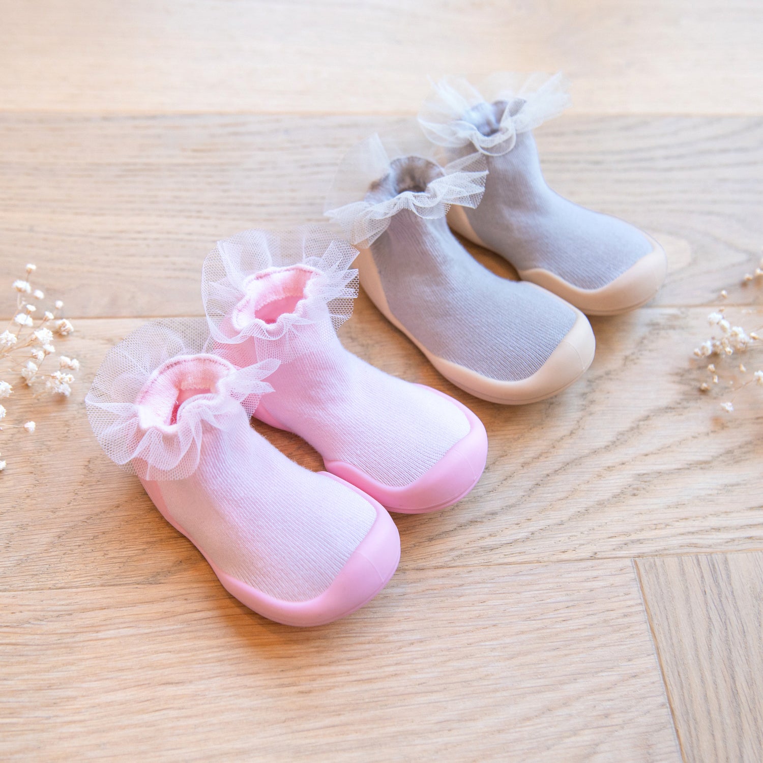 Two pairs of baby socks in pink and gray, with white lace trim, sit on a wood floor.