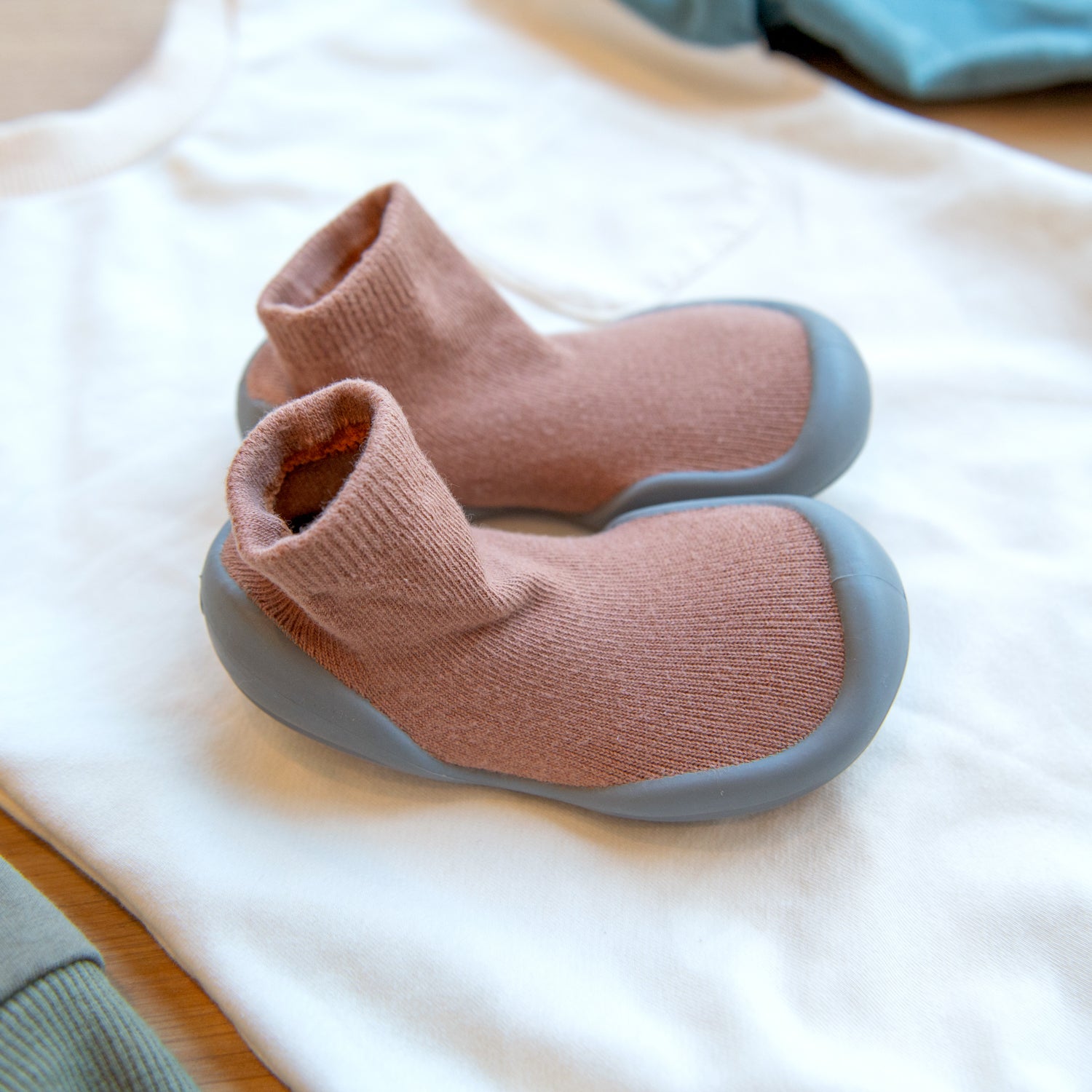 Two mauve baby sock shoes with gray soles sit on white cotton fabric.