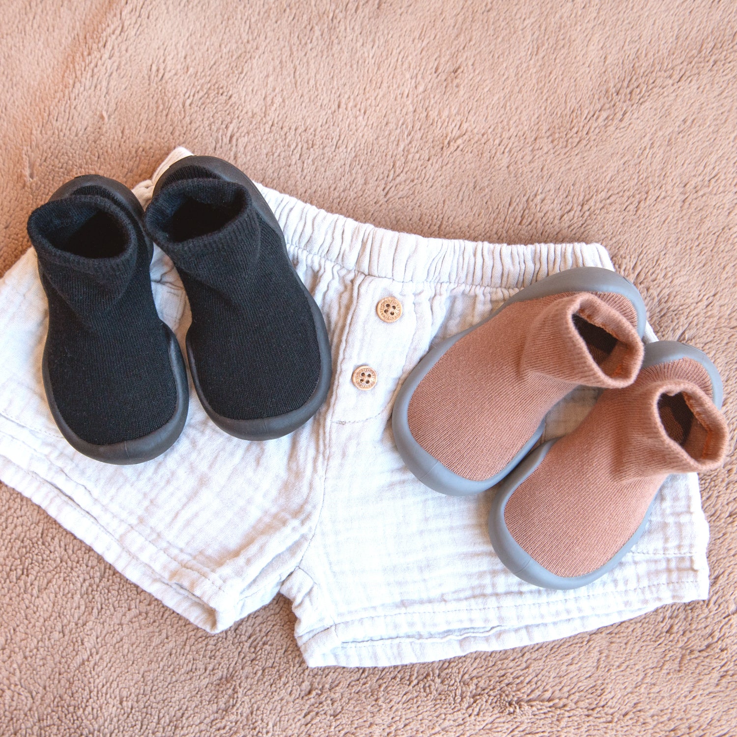 Two pairs of baby shoes and a pair of shorts on a brown rug. One pair of shoes is black, and the other is a light brown color. The shorts are white with small wooden buttons.