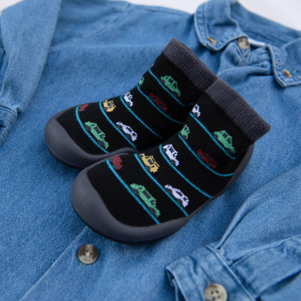 A pair of black sock shoes with colorful car designs resting on a light blue denim shirt.