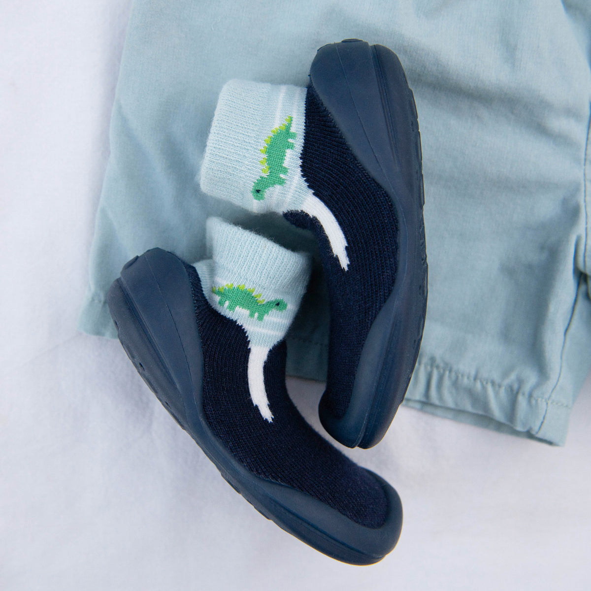 Two navy baby shoes with dinosaur socks next to light blue shorts.