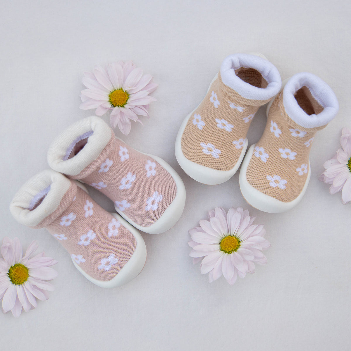 Two pairs of baby shoe-socks with flower patterns and white soles, arranged with pink daisies on a white background.