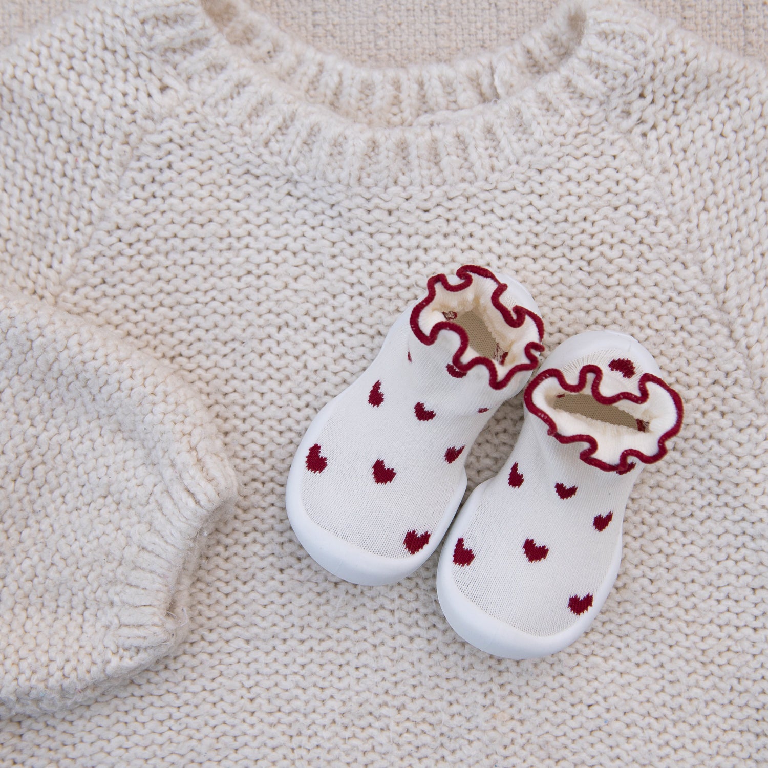White baby shoes with red heart patterns on a beige knitted surface