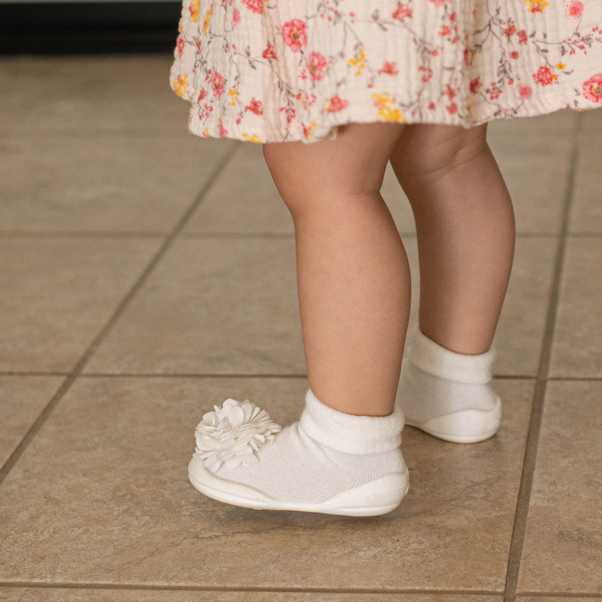 Toddler legs in white socks and shoes with a flower detail, standing on a tiled floor, wearing a floral dress.