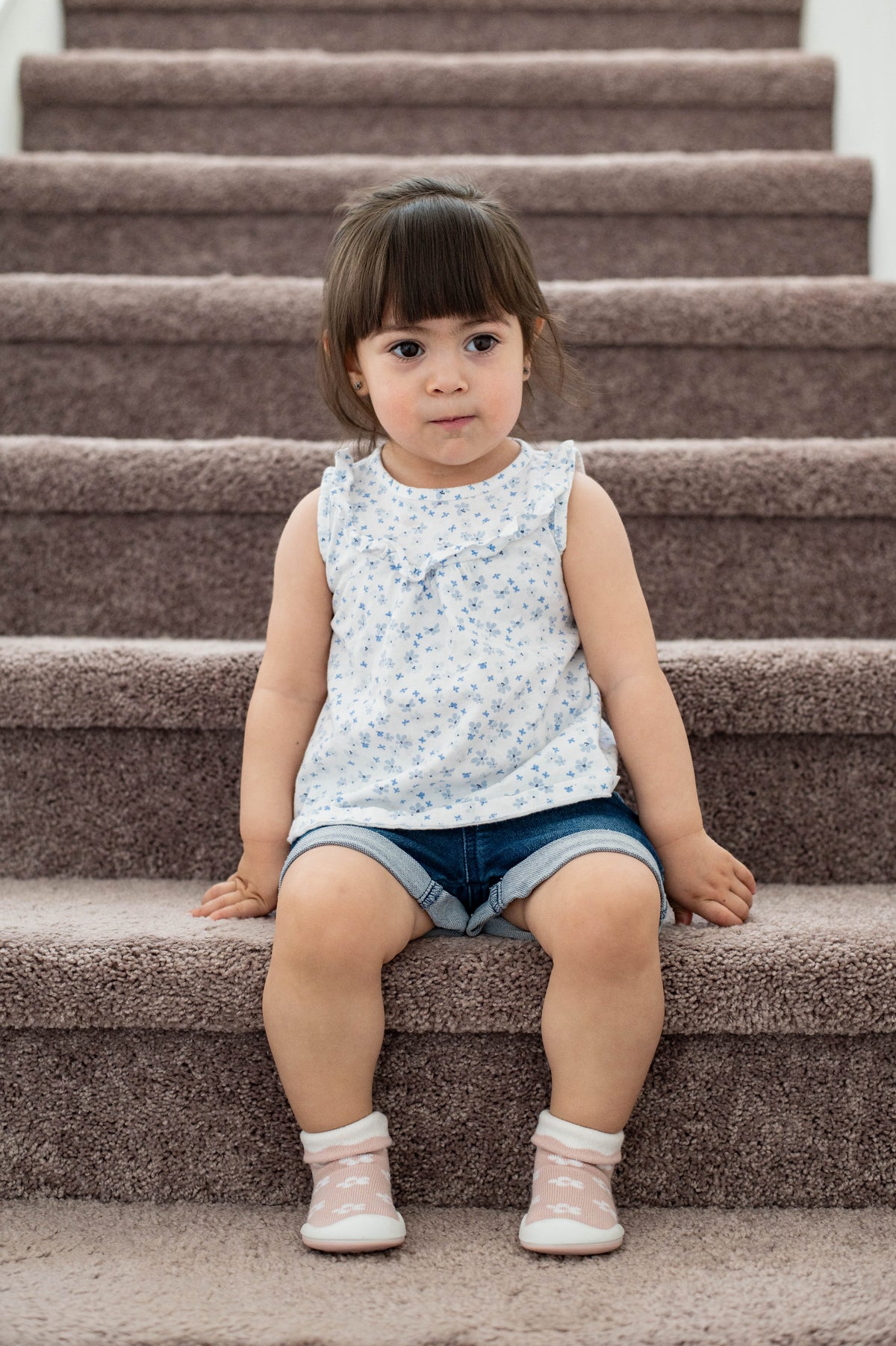 Young child sits on carpeted stairs. Sleeveless floral shirt and denim shorts. Pink and white booties.