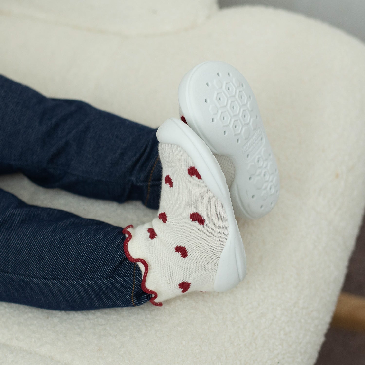 White shoes with red patterns on a child's feet wearing blue jeans, sitting on a white surface.