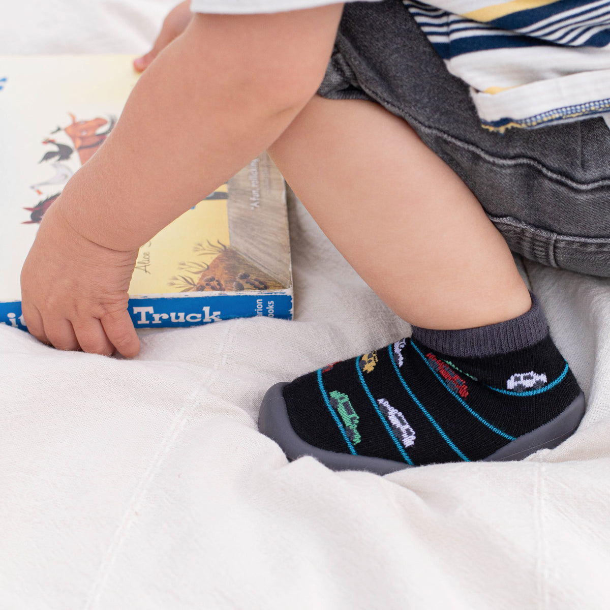 Child sitting with book, wearing gray shorts, striped shirt, and black car-patterned sock shoes.