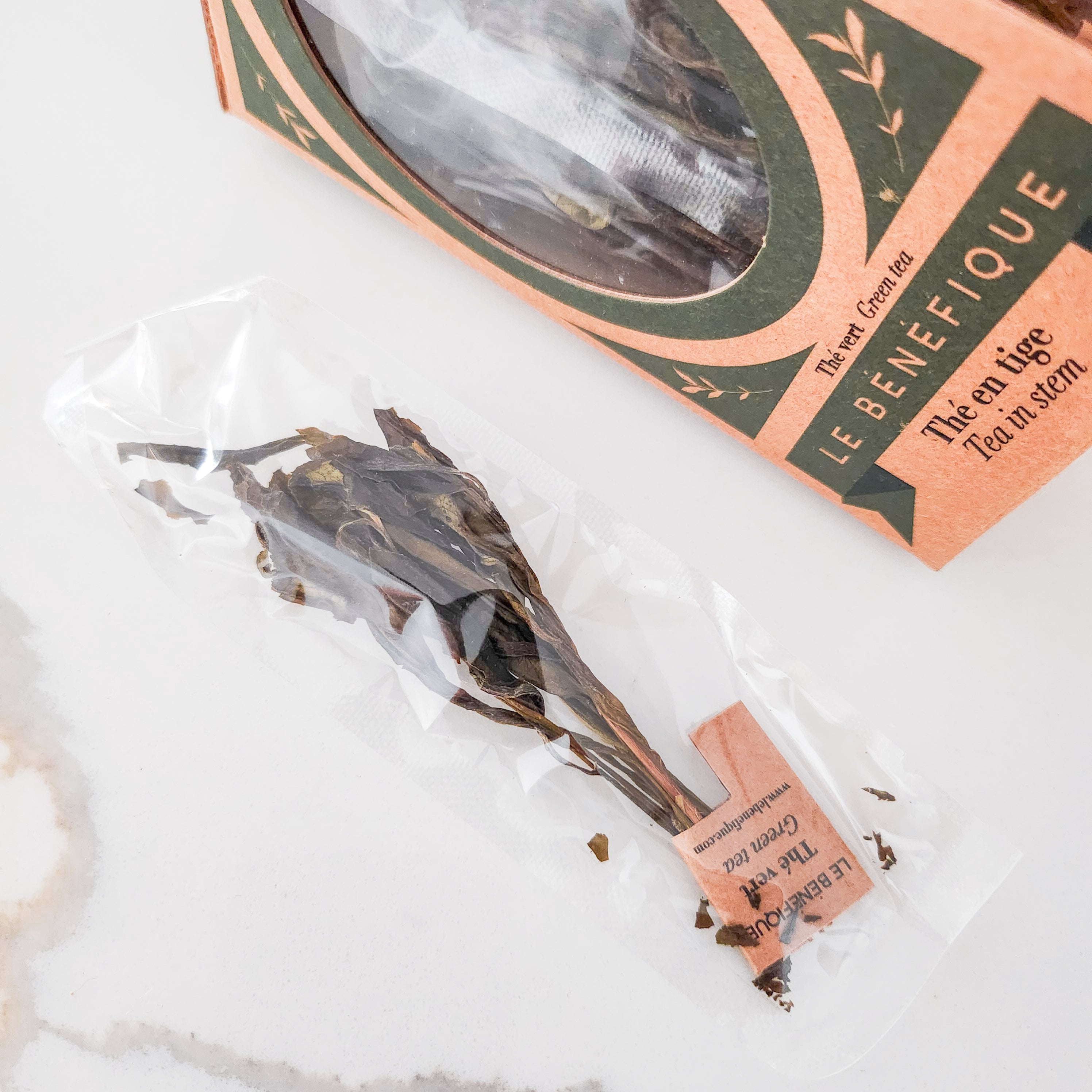 Close-up of Le Benefique Green Tea in stem, packaged in a clear plastic bag, next to a box of the same tea, on a white marble surface.